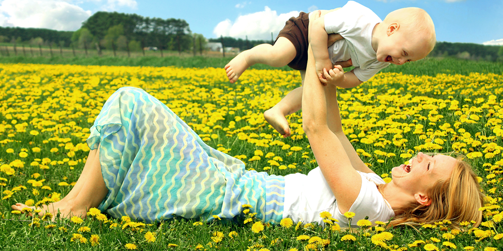 Glückliche alleinerziehende Mutter liegt in einer Blumenwiese und hält ihr lachendes Kind in die Luft